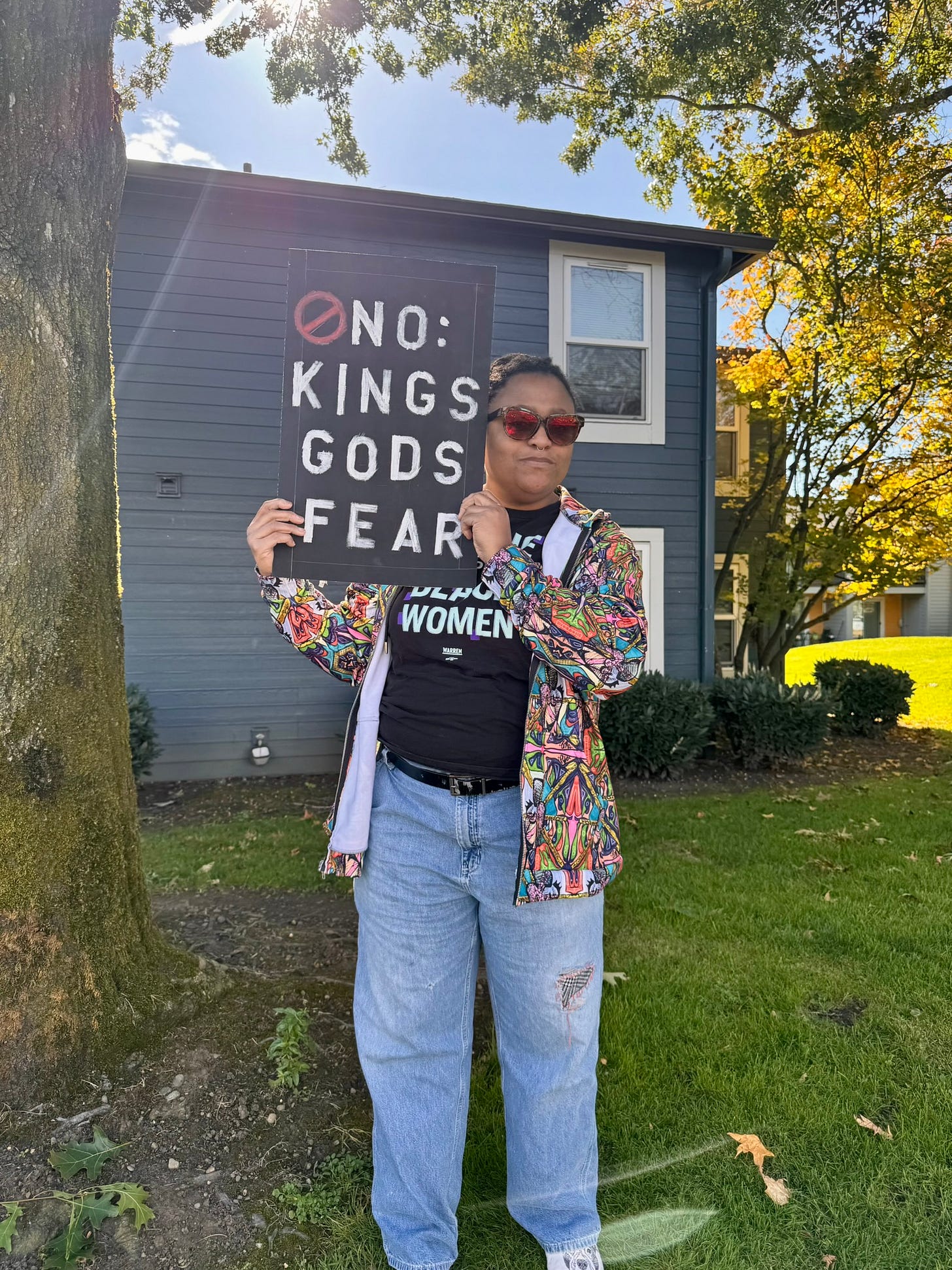 A woman stands outdoors beneath a tree, holding a black protest sign that reads “No: Kings Gods Fear.” She wears red sunglasses, jeans, and a colorful patterned jacket. Sunlight filters through the branches above her, evoking calm strength and quiet confidence. A woman stands outdoors beneath a tree, holding a black protest sign that reads “No: Kings Gods Fear.” She wears red sunglasses, jeans, and a colorful patterned jacket. Sunlight filters through the branches above her, evoking calm strength and quiet confidence.