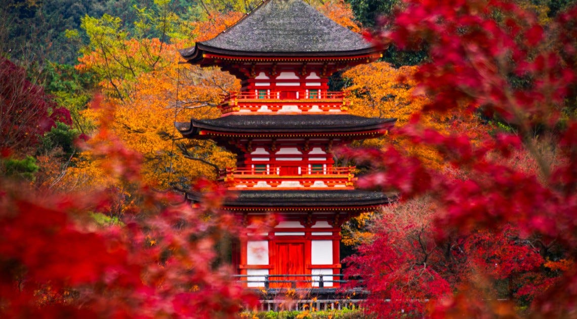 A vivid red three-story pagoda framed by autumn foliage in deep crimson, orange and gold, at Kiyomizu-dera temple in Kyoto.