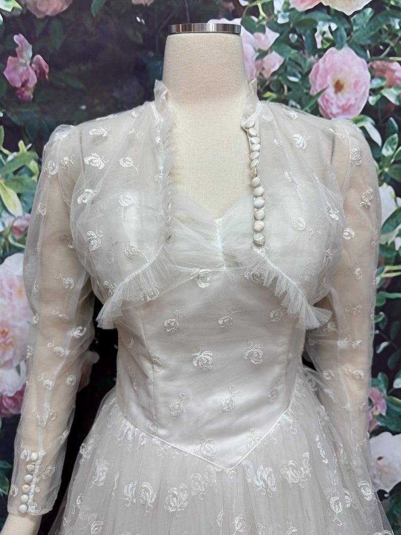 May include: A white vintage wedding dress with a lace overlay and a sweetheart neckline. The dress has a fitted bodice and a full skirt. The dress is accessorized with a pearl necklace and a white bolero jacket with lace detailing.