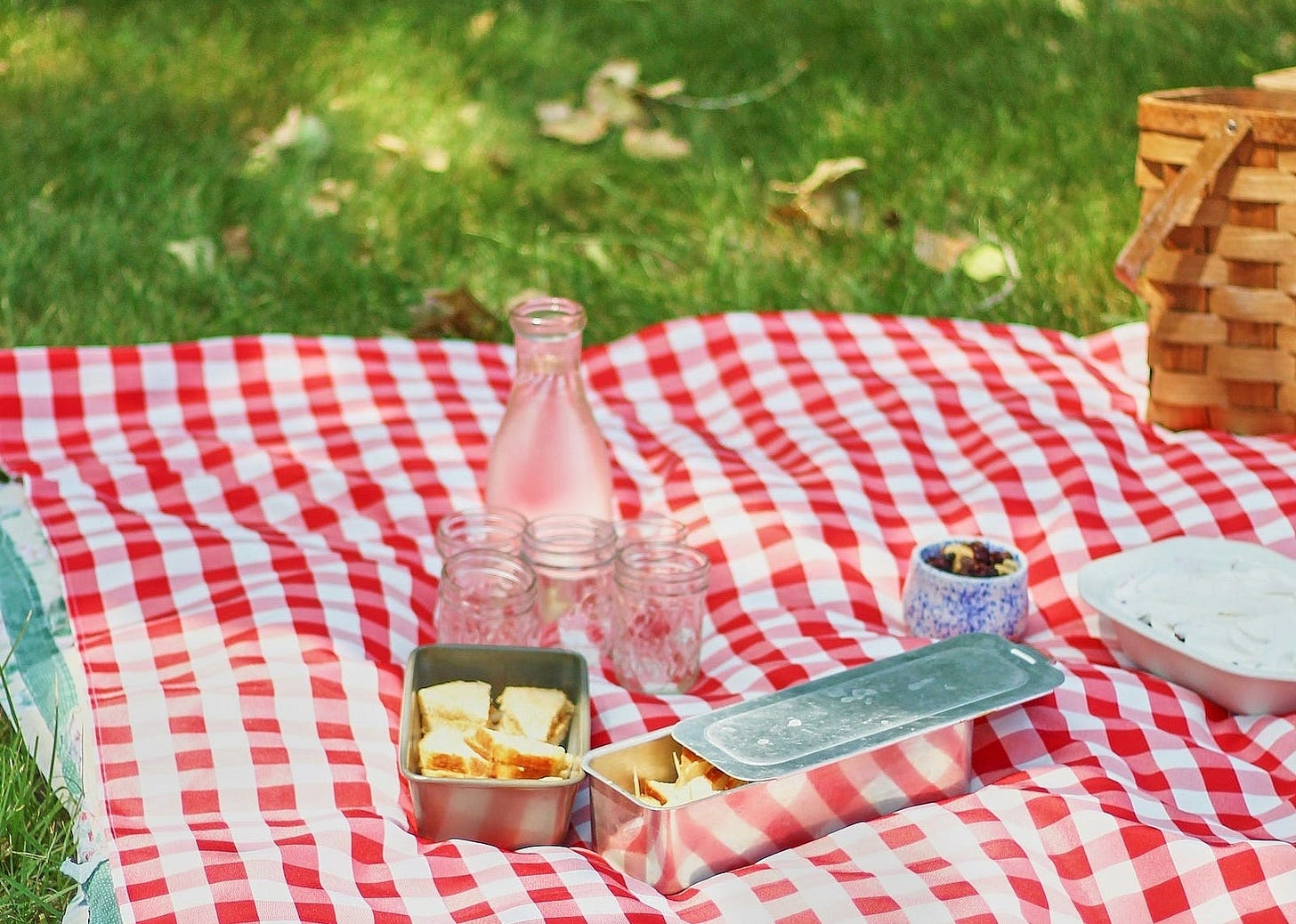 Photograph of a red and white checked picnic blanket on green grass, with dappled sunlight. There are some picnic items on the blanket and the edge of a picnic basket is visible to the far right. Photograph of a red and white checked picnic blanket on green grass, with dappled sunlight. There are some picnic items on the blanket and the edge of a picnic basket is visible to the far right.