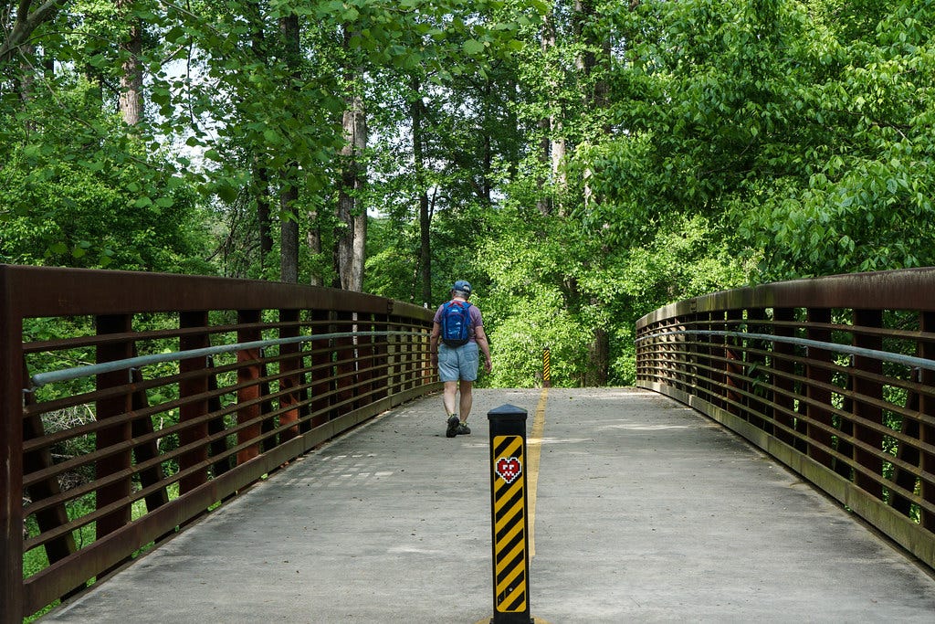 A man walking over a bridge on the Atalanta Beltline. There are a lot of green trees ahead of him as he walks away from the camera A man walking over a bridge on the Atalanta Beltline. There are a lot of green trees ahead of him as he walks away from the camera