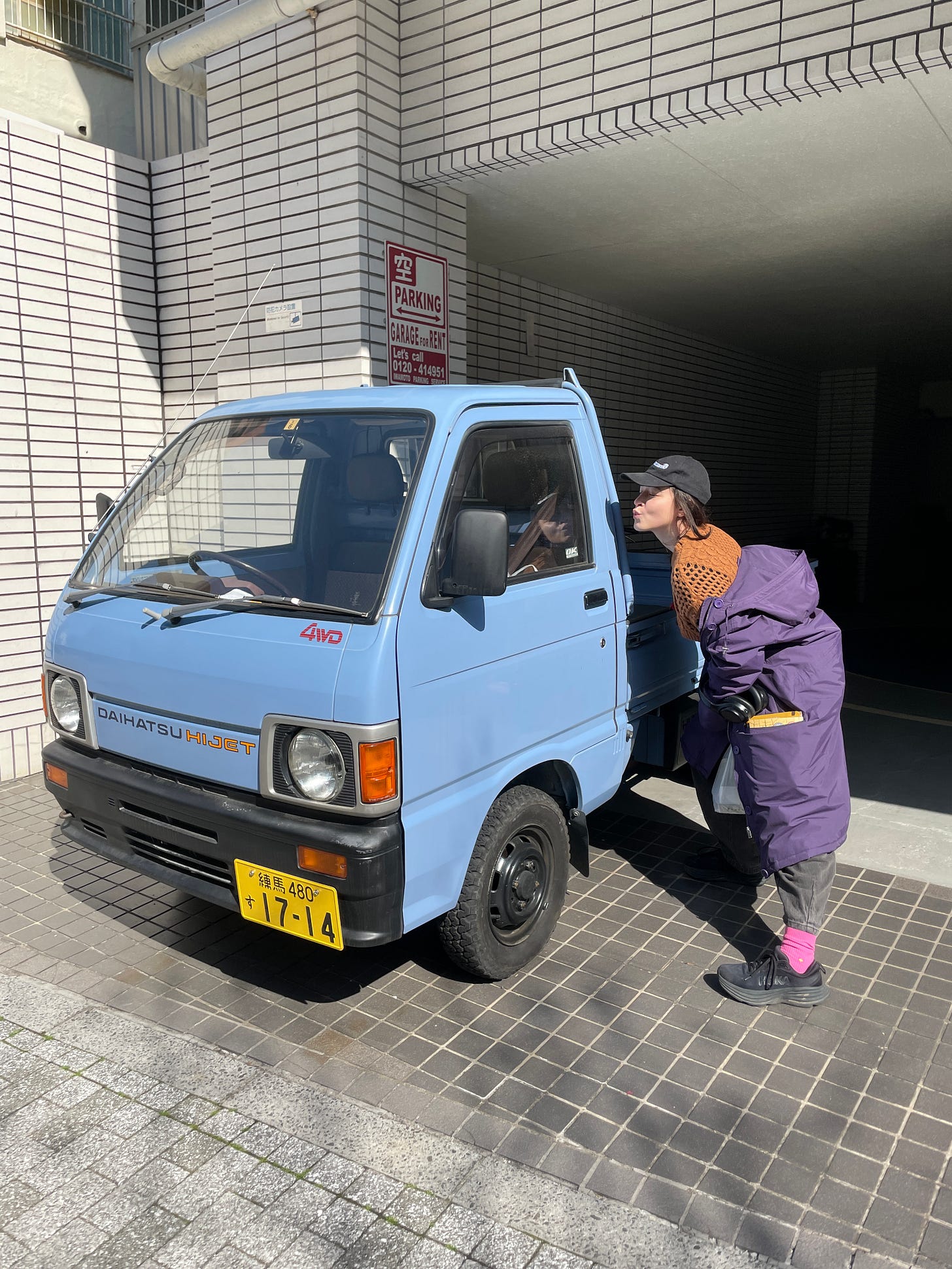 Macie poses, making a kissing face, with a powder blue mini truck in Japan.