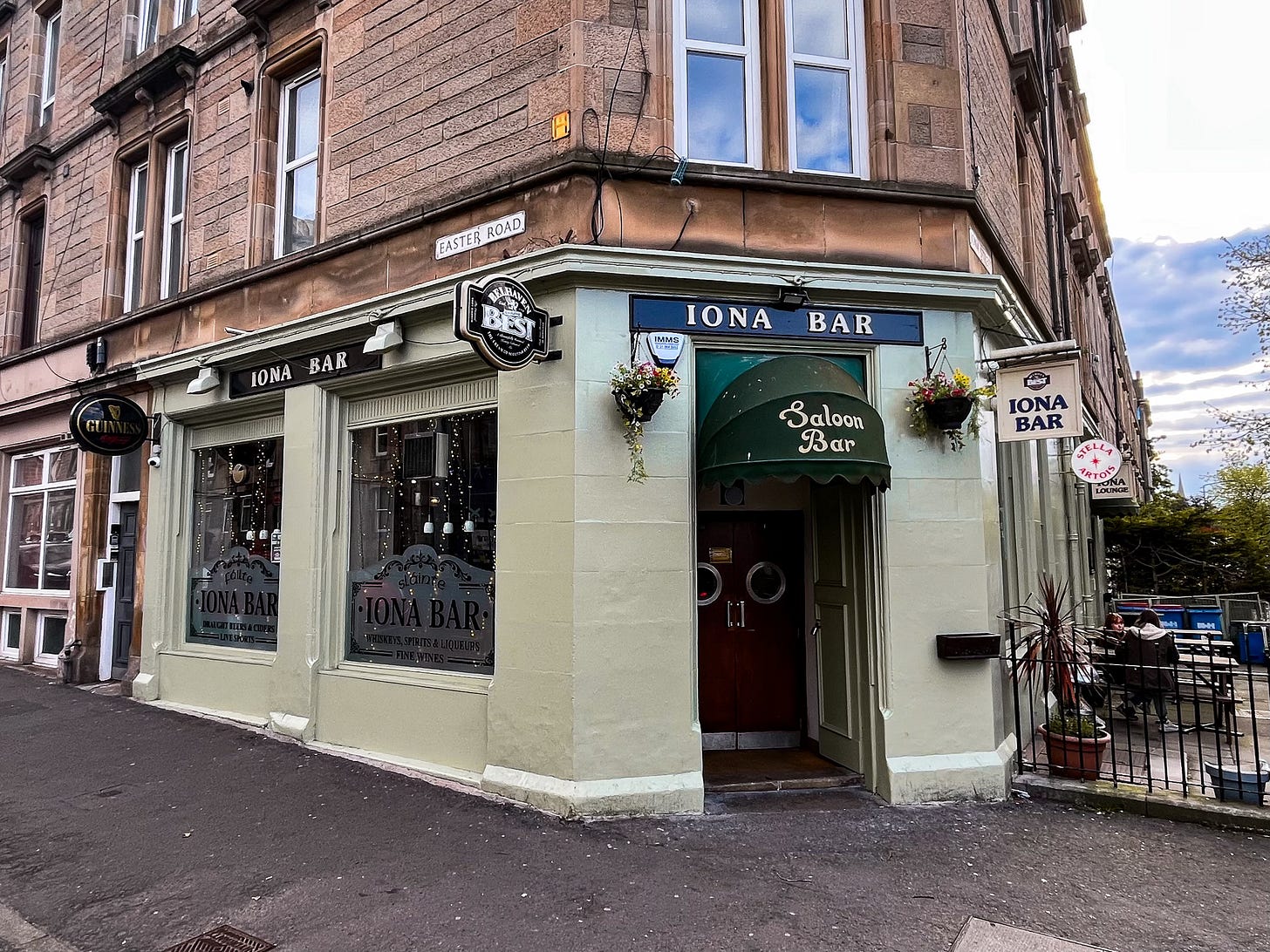 The light green exterior of Iona Bar. The sign is white writing on a black background. Guinness and Belhaven Best logos are outside.