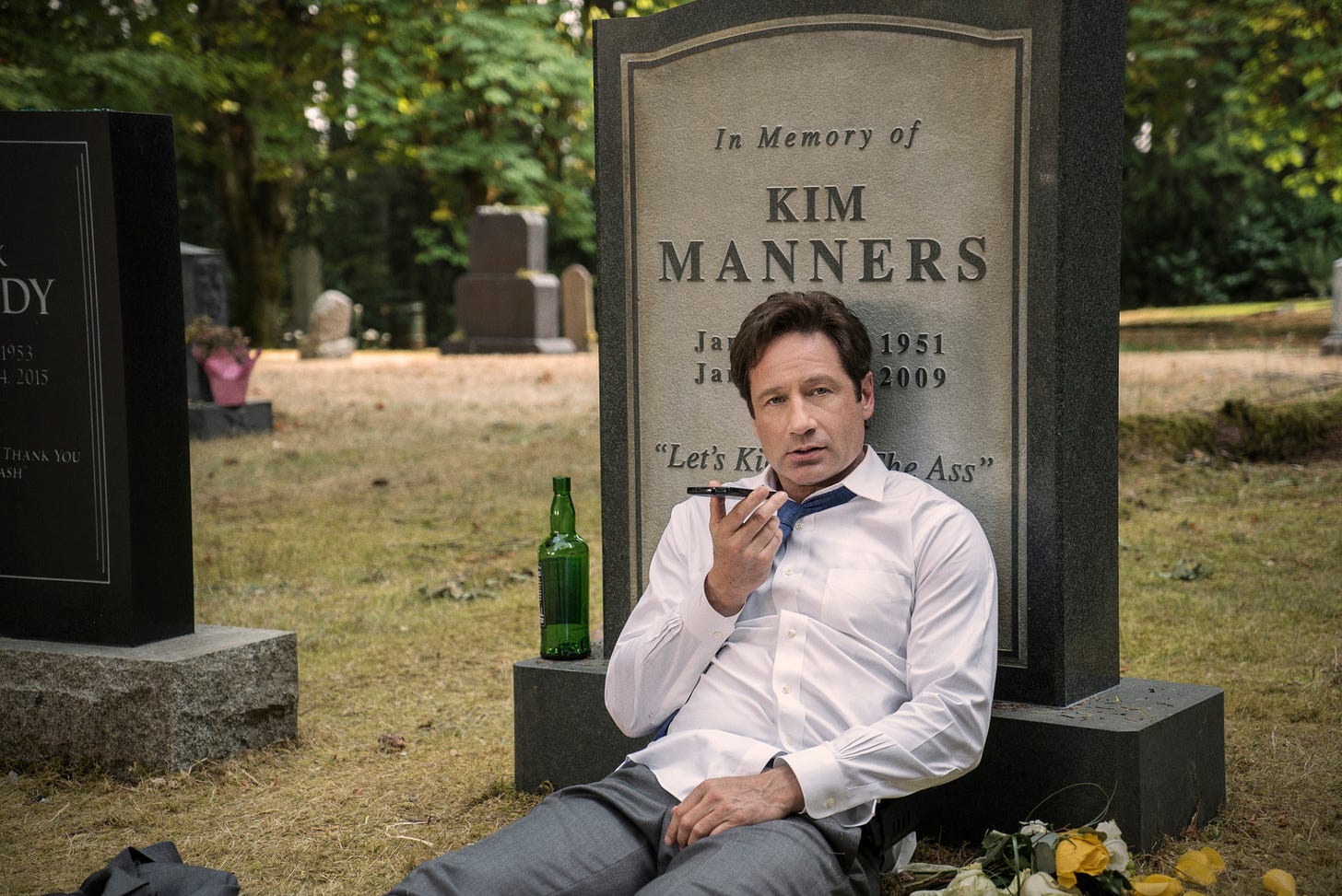 A screenshot from an episode of the 2016 reboot of the X Files. Fox Mulder is wearing a suit, a white shirt and a blue tie, and is sitting on the ground in a cemetery, leaning on a tombstone that reads “In Memory of Kim Manners.” The dates are partially obstructed by Mulder’s body, as well as a quote at the bottom, that says “Let’s kick it in the ass.” Mulder is holding a phone in front of his mouth. An empty bottle of liquor rests on the pedestal of the headstone. 