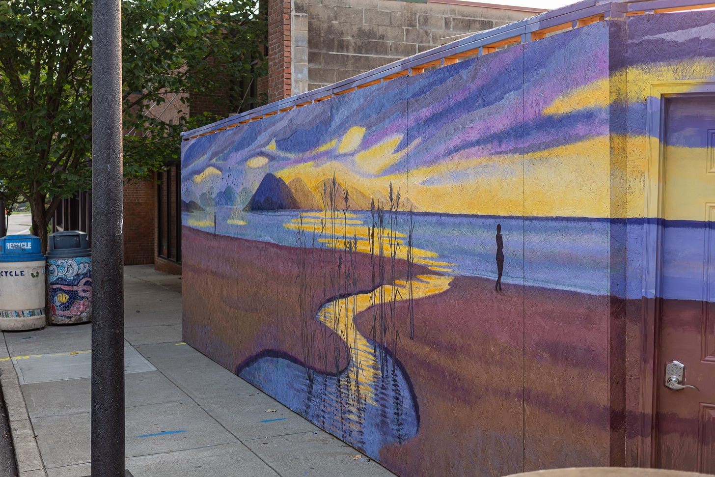 A mural of a sunset beach scene with a winding stream, warm purple and gold sky, and a lone silhouetted figure walking the shore.