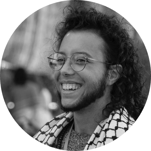 Persona con capelli lunghi ricci, sorridente, con grandi occhiali tondi. Indossa una keffiah.