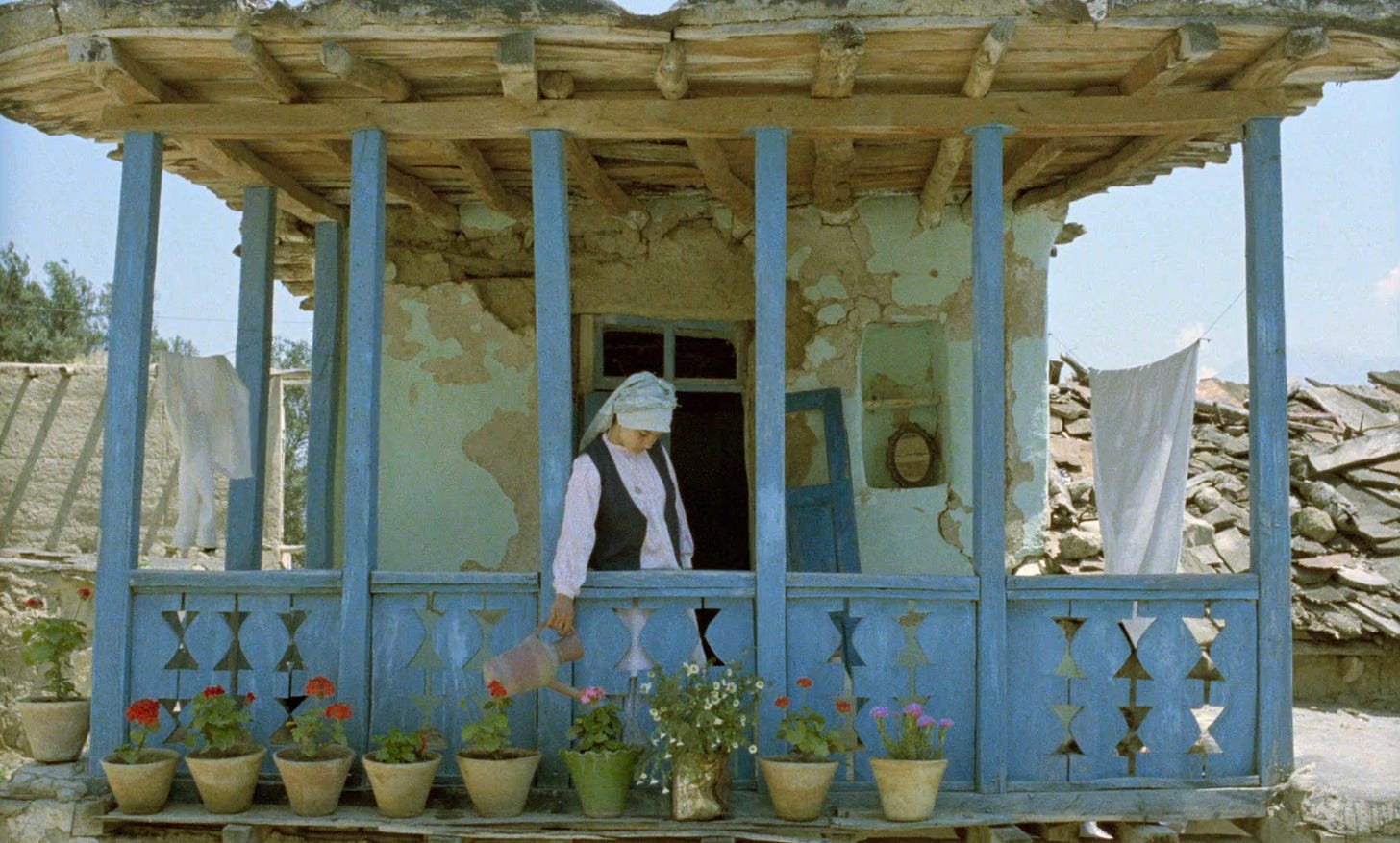 captura de tela do filme "e a vida continua", de abbas kiarostami. uma mulher rega as plantas na varanda de casa. a estrutura da casa está relativamente intacta comparada aos arredores, onde podemos ver bastante devastação. a casa em si está com as paredes fissuradas e a fachada danificada, mas segue de pé.