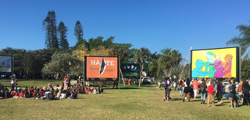 green field with trees in the background and blue skies above. In foreground are several billboards on the field, groups of people standing and sitting near the posters
