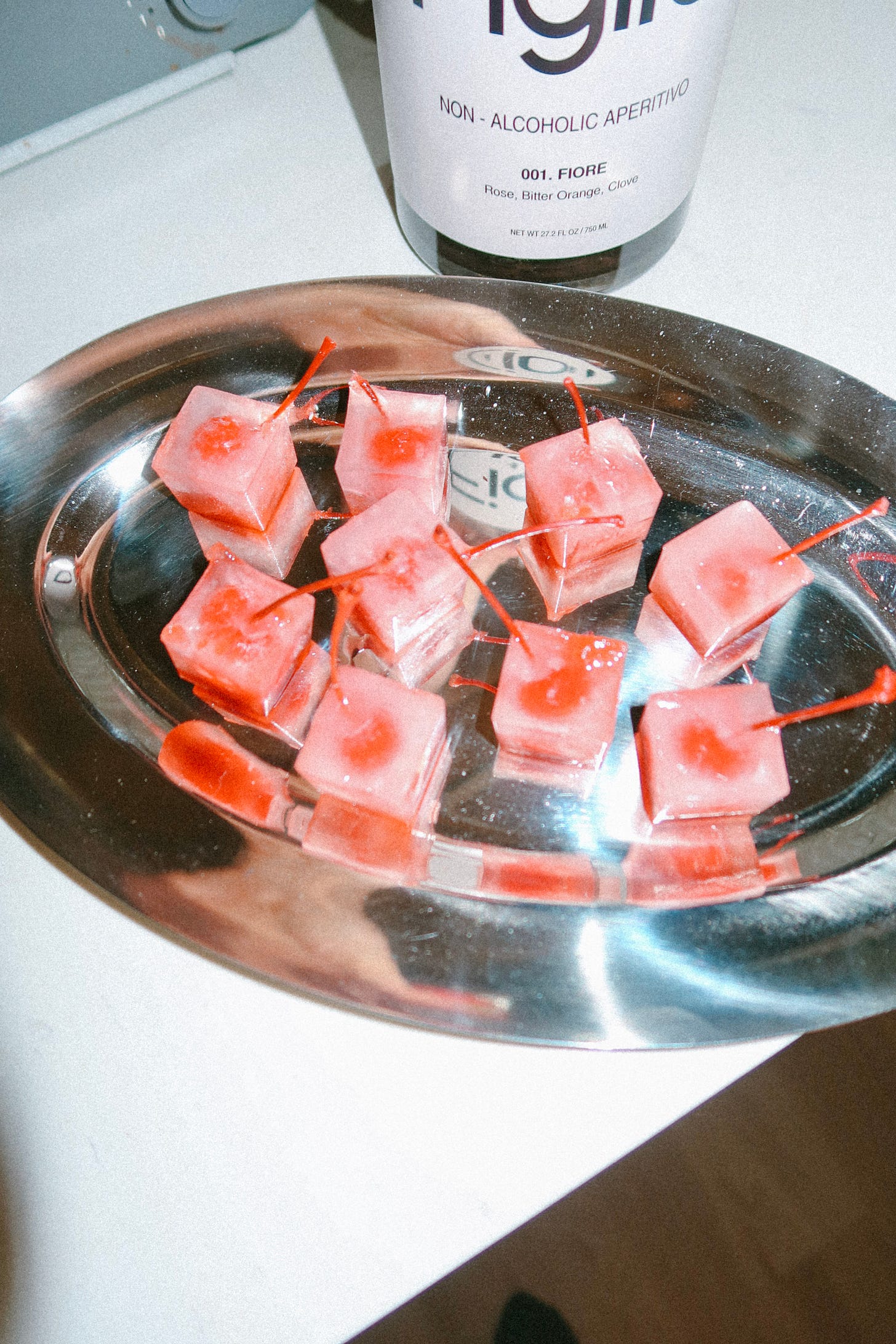 Tray of pink ice cubes topped with cherries beside a bottle of non-alcoholic aperitif