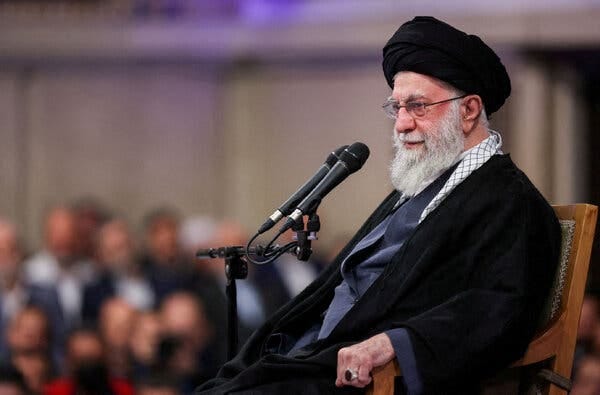 An elderly cleric with a white beard and black turban sits in a wooden chair. Two microphones are positioned in front of him.