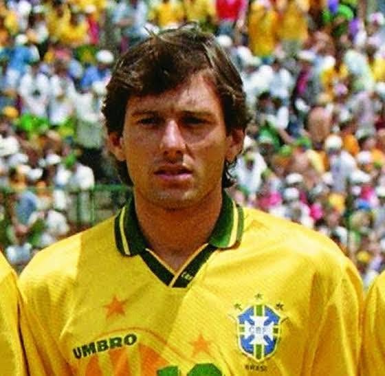 A imagem traz um retrato em plano médio do ex-jogador brasileiro Leonardo usando a camisa oficial amarela da Seleção Brasileira fabricada pela Umbro, referente à época da Copa do Mundo de 1994, com destaque para a gola polo verde e o escudo da CBF acompanhado das três estrelas do tricampeonato. Ele aparece com uma expressão séria e focada, olhando diretamente para a frente, exibindo o cabelo castanho levemente ondulado em um corte característico dos anos 90, enquanto ao fundo é possível observar uma arquibancada lotada e desfocada de torcedores vestindo cores vibrantes sob a luz natural de um estádio de futebol.
