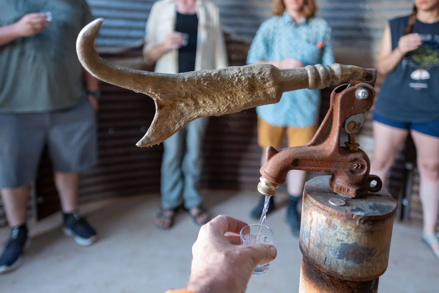 A hand reaches to fill s mall cup with water pouring from "The Tap," an artwork by M12 that features a found antler handle to a tap. A group of people holding small cups stand in the backgrounc, which is blurred. A hand reaches to fill s mall cup with water pouring from "The Tap," an artwork by M12 that features a found antler handle to a tap. A group of people holding small cups stand in the backgrounc, which is blurred.