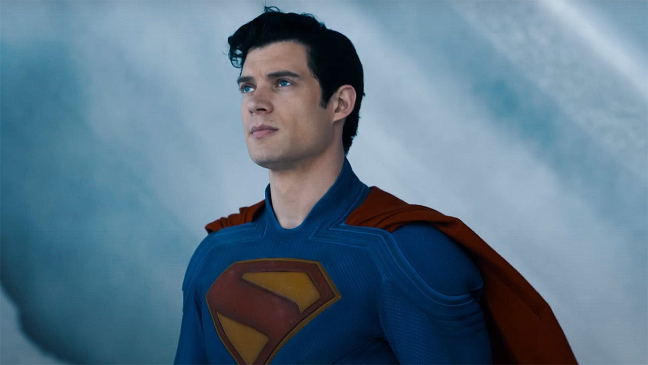 David Corensweat como Superman; ele é um homem branco, de porte musculoso, cabelos pretos e olhos azuis, vestindo um macacão inteiriço de corpo todo azul, com capa vermelha, e logo amarelo e vermelho com um S de Superman.