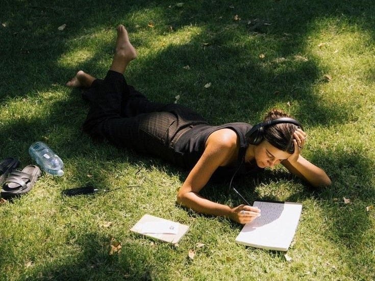 This may contain: a woman laying on the grass writing in her notebook and listening to earbuds
