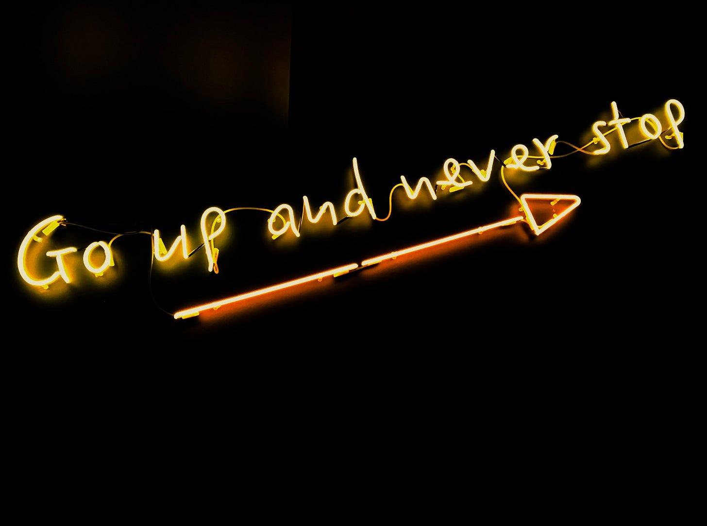 Image of yellow neon lights that say, "Go up and never stop", with an arrow pointing upwards.