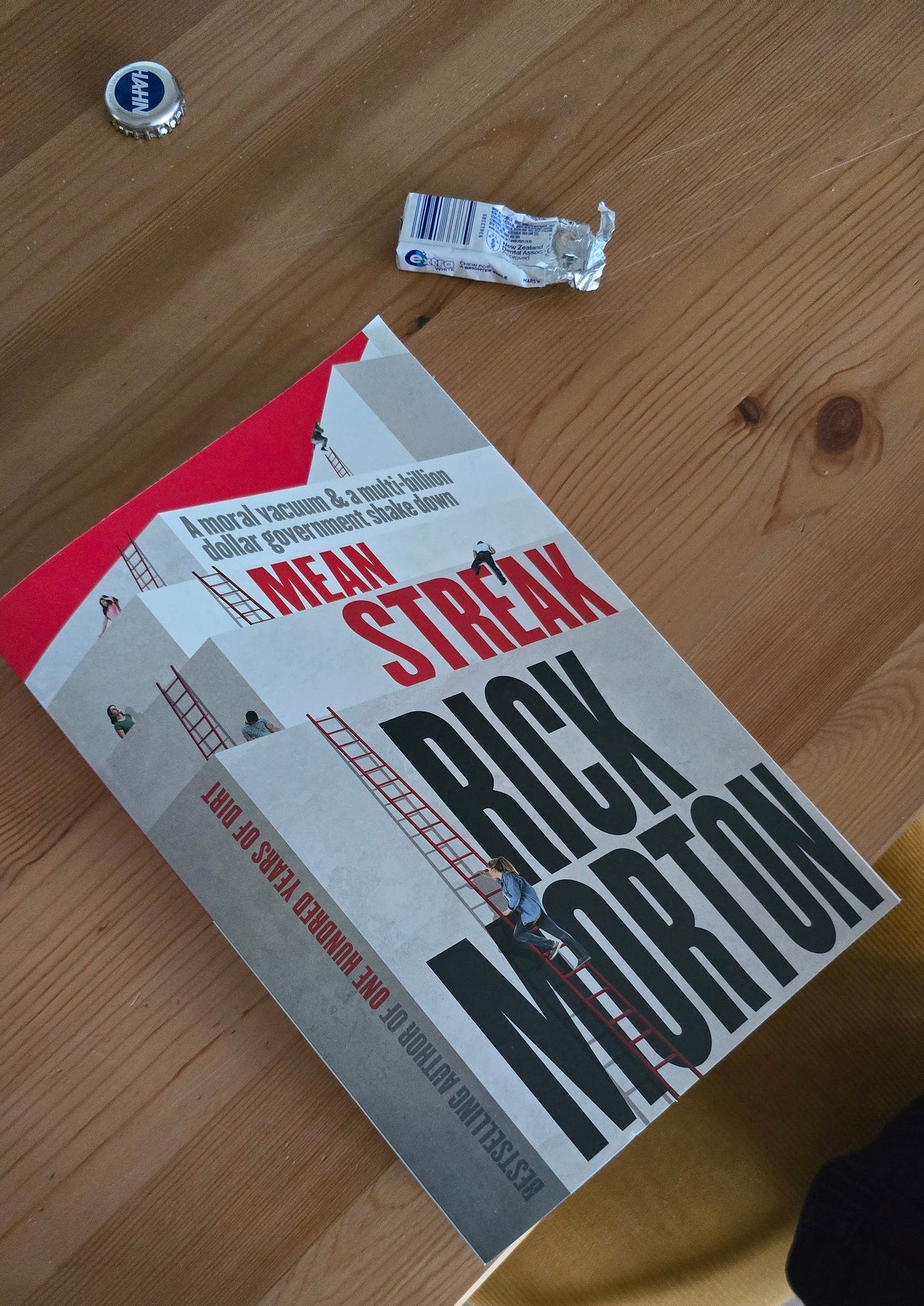 It's a photo of the book 'Mean Streak' by Rick Morton, placed on a wooden table. Slightly above it are a bottle cap for HAHN Superdry and half a packet of Extra Whitening Gum. 