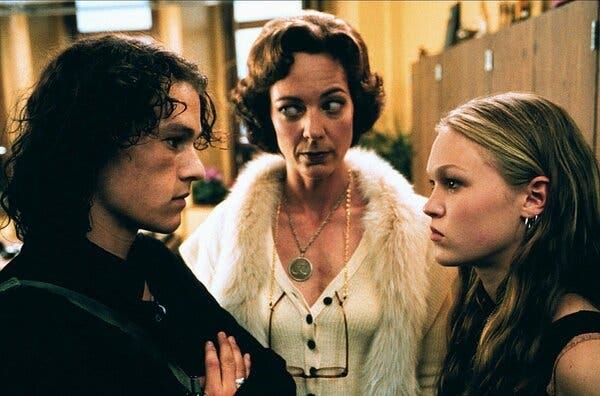 A scene from the movie “10 Things I Hate About You.” Heath Ledger, with long black hair, stares down Julia Stiles, with long dirty, blonde hair. Allison Janney, wearing a white blouse and white boa, stands in between them.