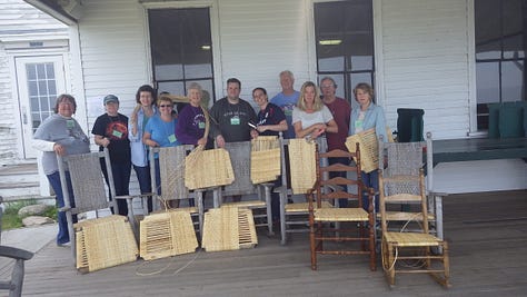A rocker with a finished caned back, a group of caners show off their work, Jo in a blue t-shirt sitting in a finished rocker.