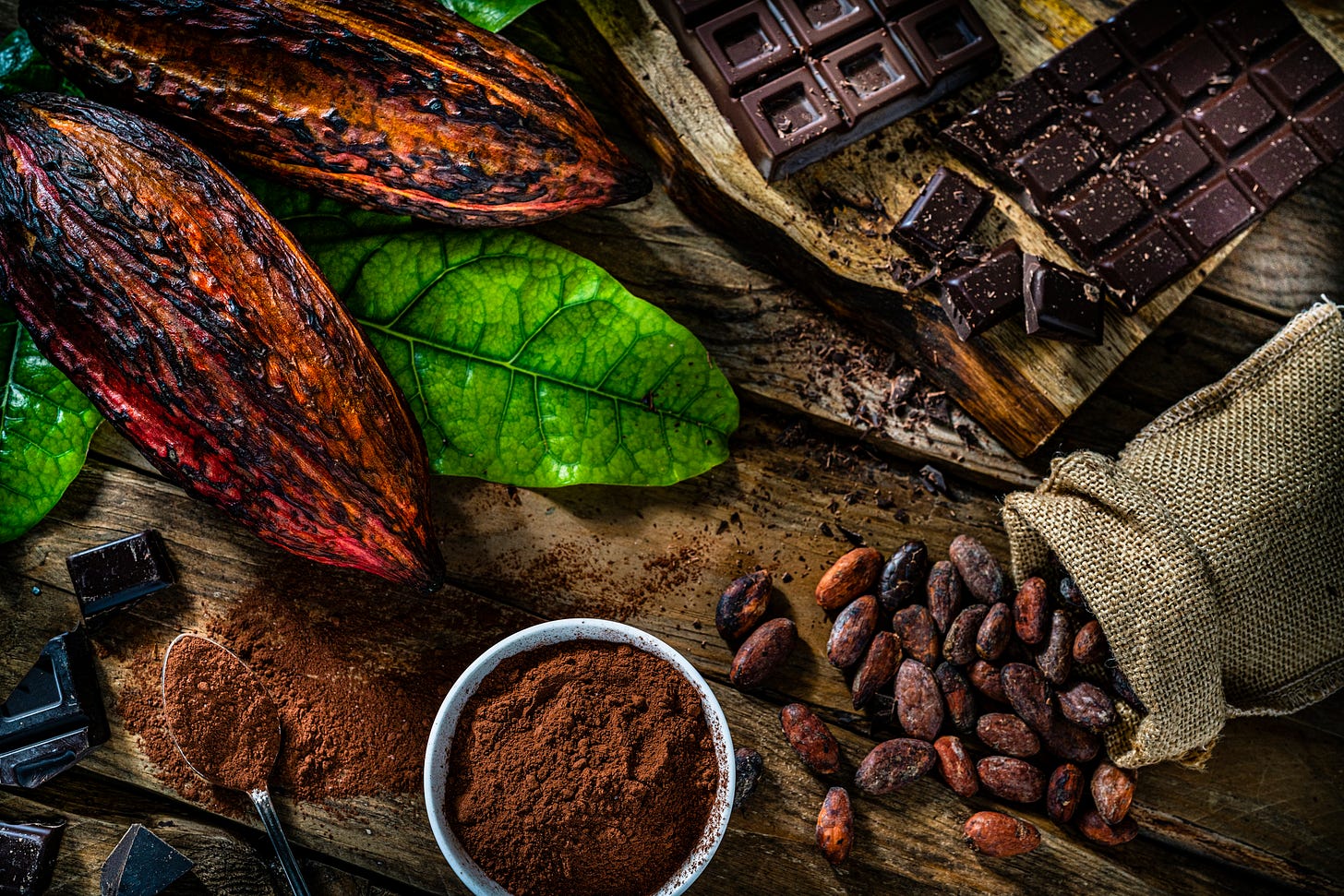 A rustic wooden table displays elements of chocolate production: two large cacao pods, green leaves, a bowl of cocoa powder, a burlap sack spilling cacao beans, and scattered pieces of dark chocolate bars and broken chocolate