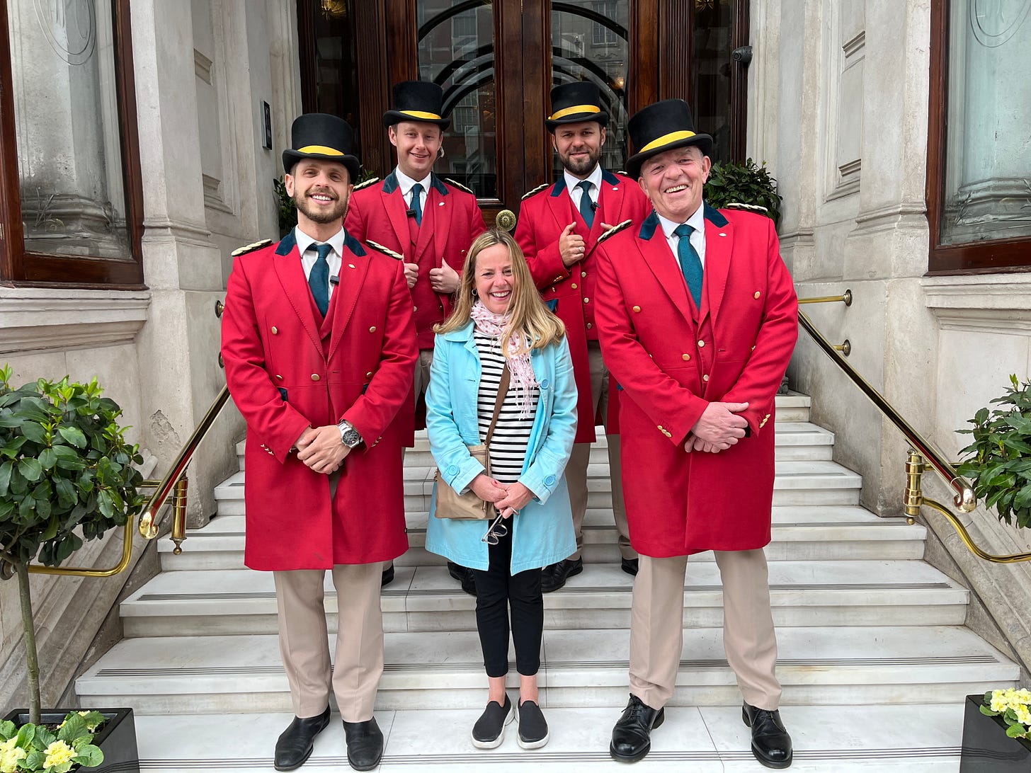 A short, blonde woman in a blue coat and pink and white polkadot scarf stands on a set of cement stairs, surrounded by four tall men in long red coats and black top hats.