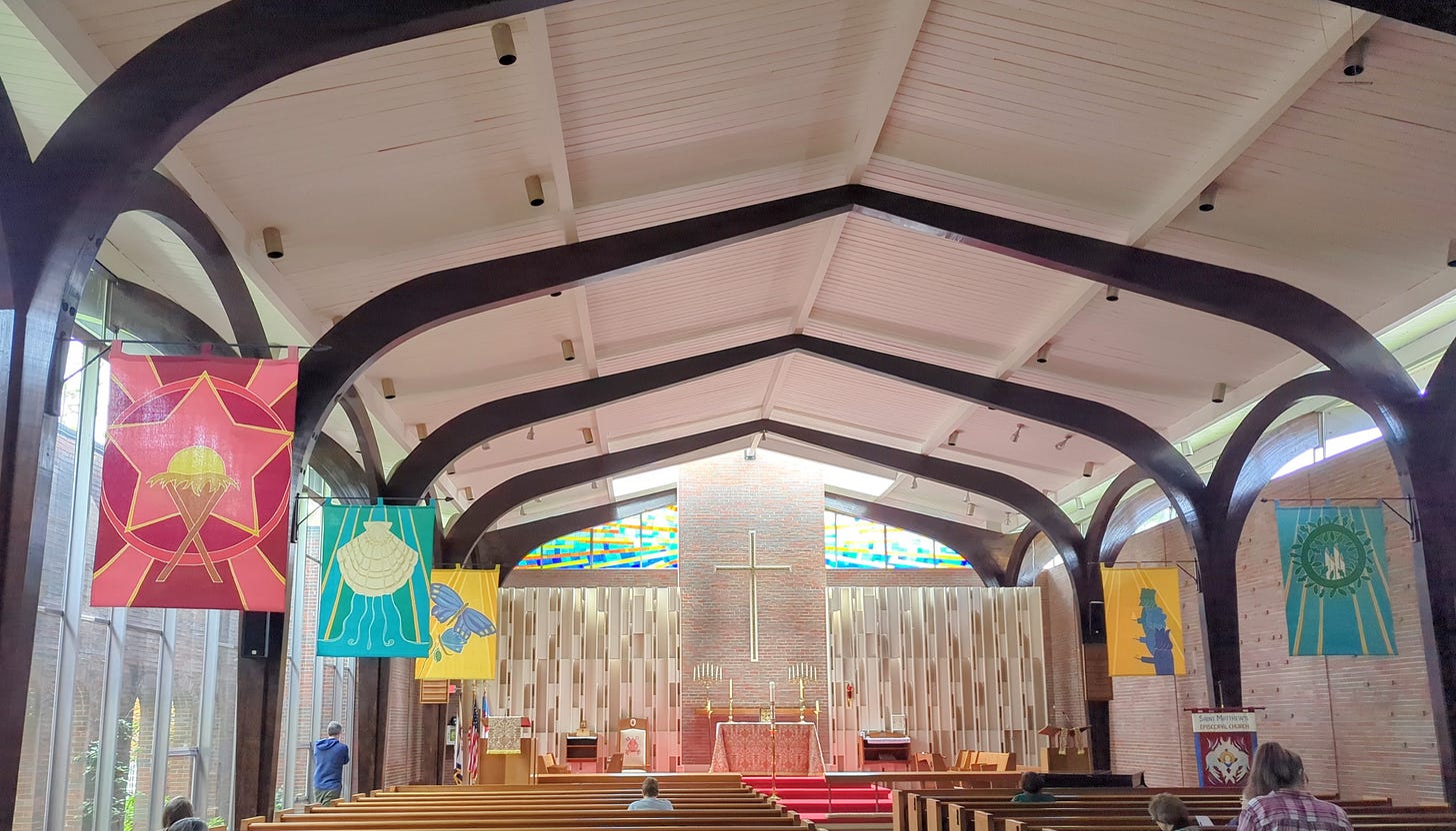 The inside of a bright sanctuary. The wall on the left is all glass and there is stained glass behind the altar. Brightly colored banners project out into the space. 