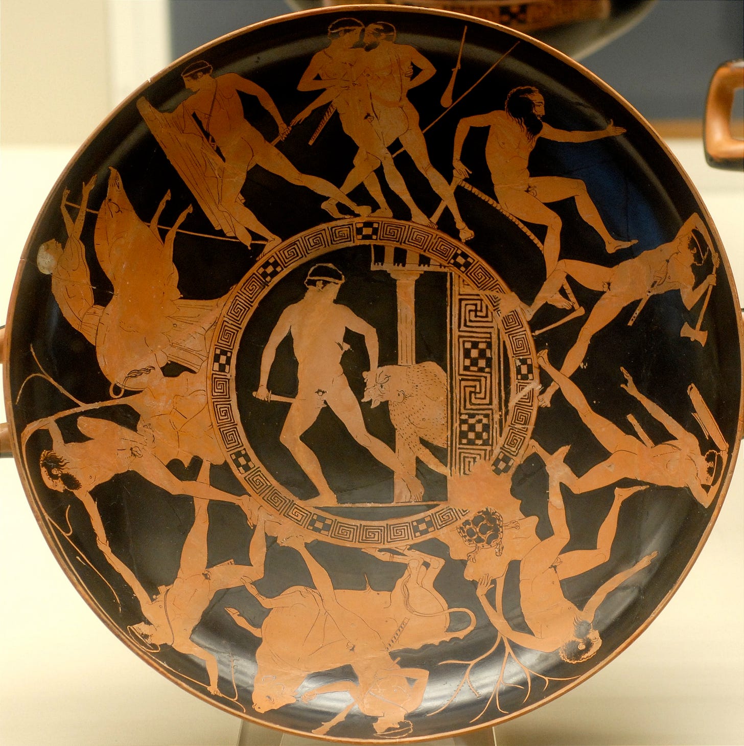 An ancient Greek red-figure kylix interior showing the Labors of Theseus. The central figure of Theseus stands with a club, flanked by the Marathonian Bull, while around the border are scenes of his various encounters, including battles with bandits and mythological creatures, all framed by a decorative meander pattern.
