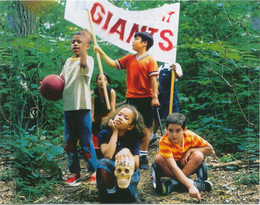 Children in a forest holding a homemade “GIANTS” banner, with one child clutching a red ball and another resting their chin on a plastic skull labeled “THEY.”