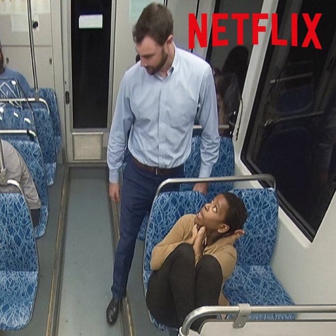 A man in a light blue shirt and dark pants stands on a bus, holding onto a pole. A woman in a brown top and black pants sits on a blue-patterned seat, looking up at the man. The bus has blue-patterned seats and metal handrails. The word "NETFLIX" in red text is overlaid at the top of the image.