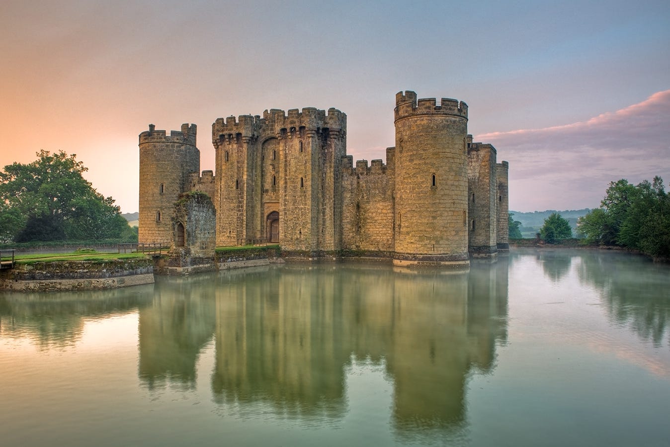 Photo of Bodiam Castle at sunset with towers and battlements reflected in a wide moat. A path leads away from the entrance of the castle across two islands Photo of Bodiam Castle at sunset with towers and battlements reflected in a wide moat. A path leads away from the entrance of the castle across two islands