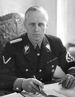 Portrait of a middle-aged man with short grey hair and a stern expression. He wears a dark military uniform, with a swastika on one arm. He is seated with his hands on a table with several papers on it, holding a pen. Portrait of a middle-aged man with short grey hair and a stern expression. He wears a dark military uniform, with a swastika on one arm. He is seated with his hands on a table with several papers on it, holding a pen.