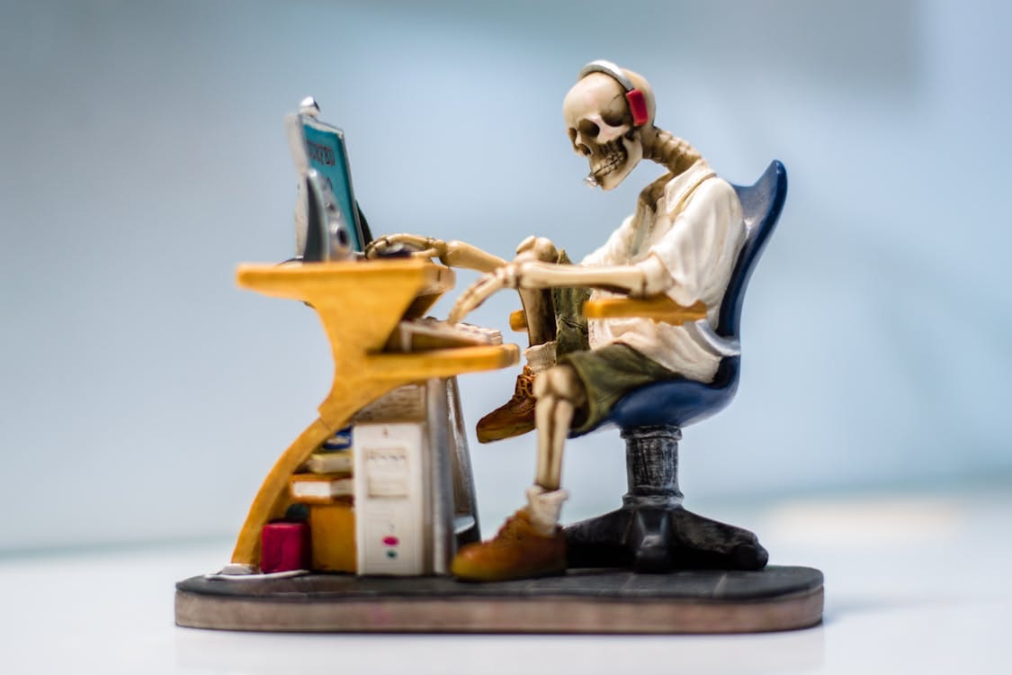 Free A quirky skeleton figurine wearing headphones sits at a desk with a computer, symbolizing burnout. Stock Photo