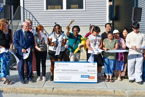Photo of elected officials and Habitat homebuyers during a ribbon cutting, holding a large symbolic check from the City of Albany and a photo of staff from Community Preservation Corporation in front of Habitat homes.