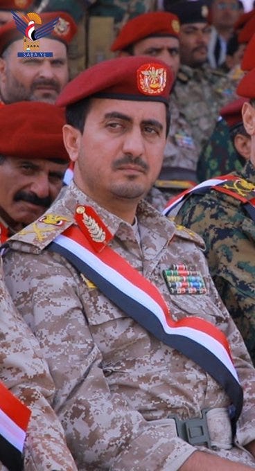Group of men in military uniforms including camouflage fatigues with red and white sash, red berets adorned with emblems, and insignia on shoulders, seated in rows at a formal gathering, central figure in foreground wearing red beret and camouflage with colorful ribbons and medals.