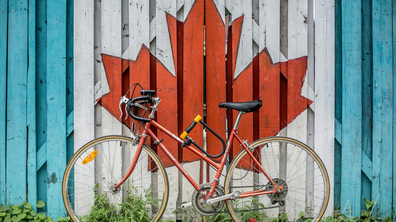 A bicycle leans against a weathered wooden fence, painted with center portion of the Canadian flag (a red maple leaf on a white field). Image credit: Ali Tawfiq/Unsplash
