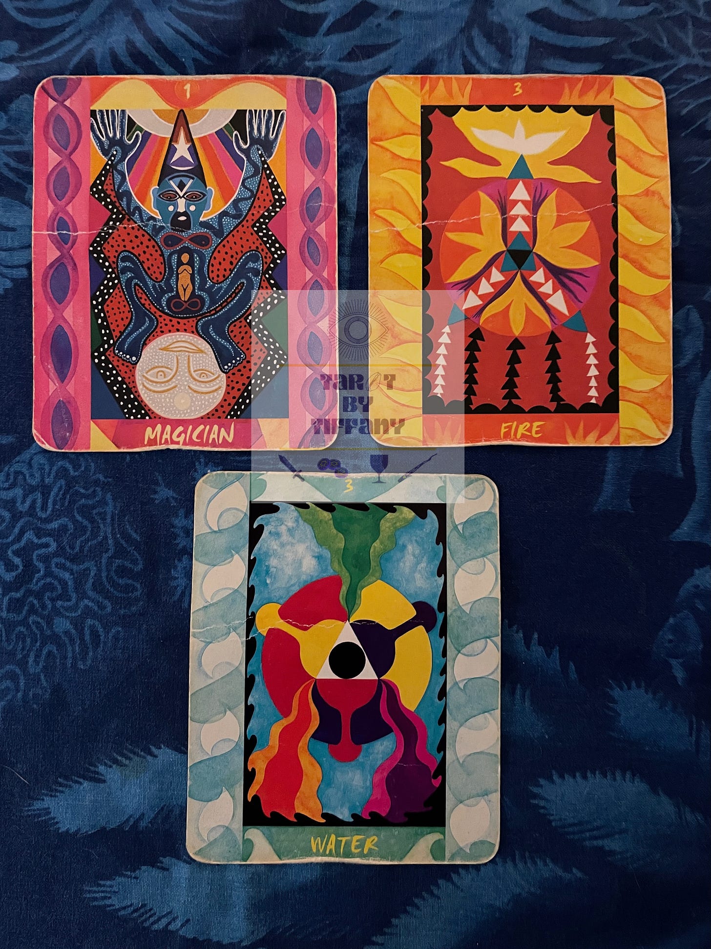 3-card reading with the Tarot of the Four Elements. The cards are laid against a cobalt blue cloth background in an aquatic motif drawn in light blue. Top two cards (L-R): I - Magician, 3 of Fire. Bottom card: 3 of Water.
