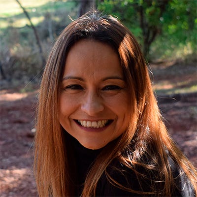 Fotografia em primeiro plano de uma mulher de cabelos castanhos claros e lisos, sorrindo abertamente para a câmera. Ela usa uma blusa preta de gola alta. O fundo é ao ar livre, desfocado, com vegetação e solo iluminados pela luz do sol Fotografia em primeiro plano de uma mulher de cabelos castanhos claros e lisos, sorrindo abertamente para a câmera. Ela usa uma blusa preta de gola alta. O fundo é ao ar livre, desfocado, com vegetação e solo iluminados pela luz do sol