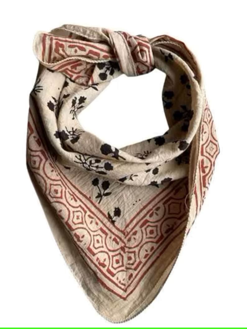 May include: A beige cotton bandana with a red and black floral print. The bandana has a geometric border and features a repeating pattern of stylized flowers and geometric shapes. The fabric appears soft and lightweight, suitable for various uses, such as a hair accessory, neck scarf, or decorative element.