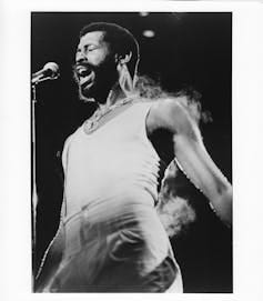 Black-and-white photo of singer wearing white undershirt singing in front of microphone, with steam coming off his body