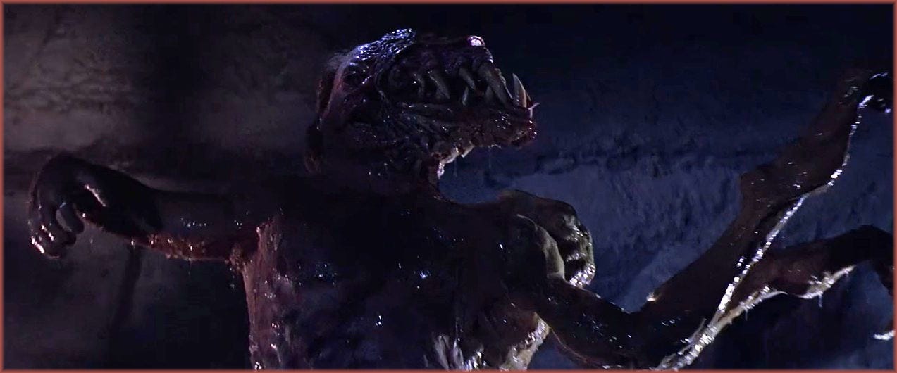 Film frame from The Thing (1982): the Blair-Monster in the underground sequence, a massive creature covered in dark rubber skin, a secondary toothed mouth growing from the side of its head, two insectoid arms extended. The creature was operated by sixty-three technicians simultaneously, with Bottin himself inside controlling the dog appendage.
