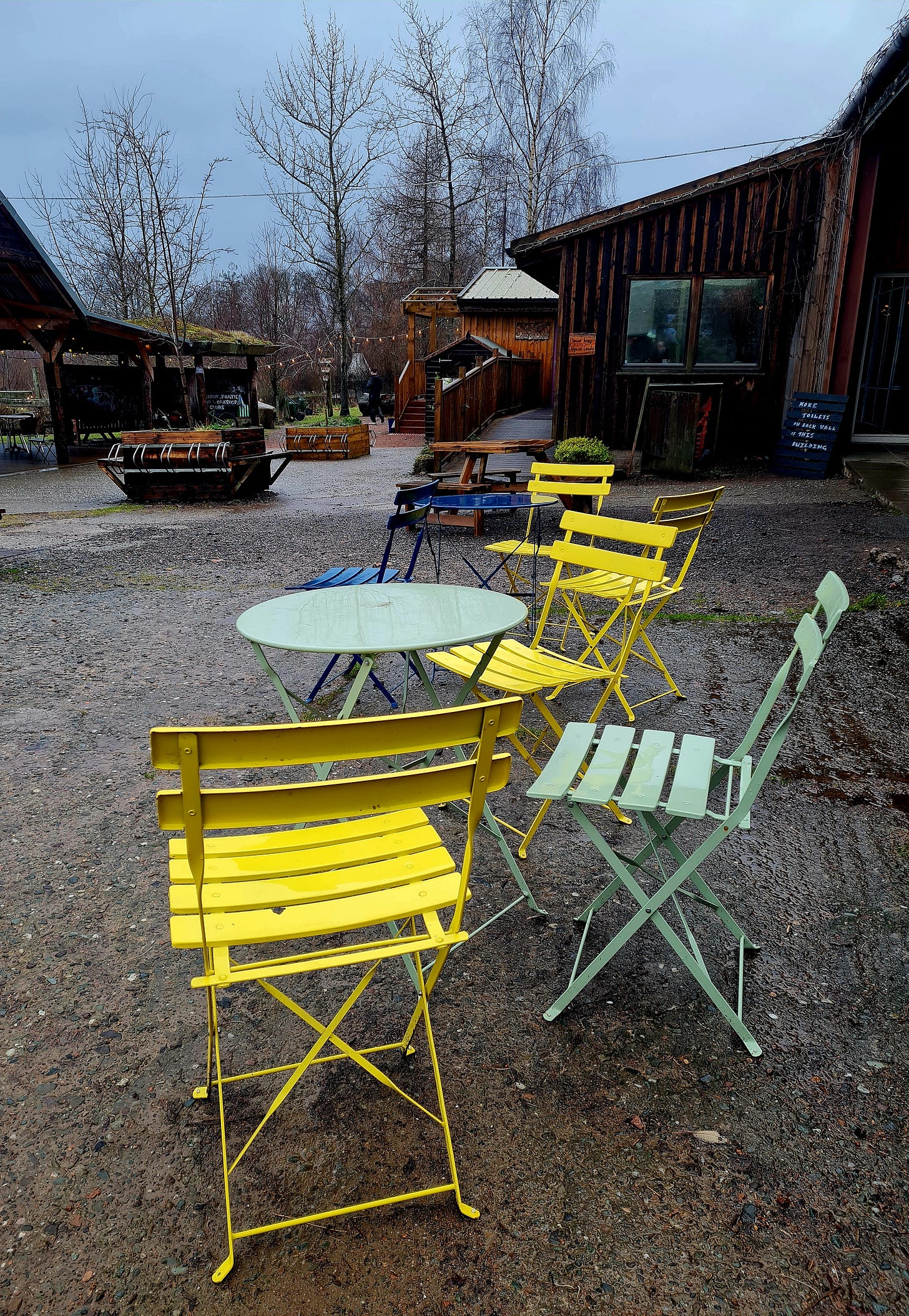 Colourful metal chairs and tables arranged outside a wooden cafe building. The weather is grey and wet, in contrast to the colourful empty seats.