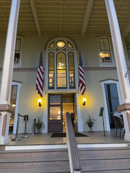 Close-up view of a Victorian hotel entrance with a tall, ornate arched window above the main door. Two American flags flank the entrance, and golden wall sconces cast a warm glow on the white clapboard exterior. The entrance includes steps with handrails and potted plants on the porch