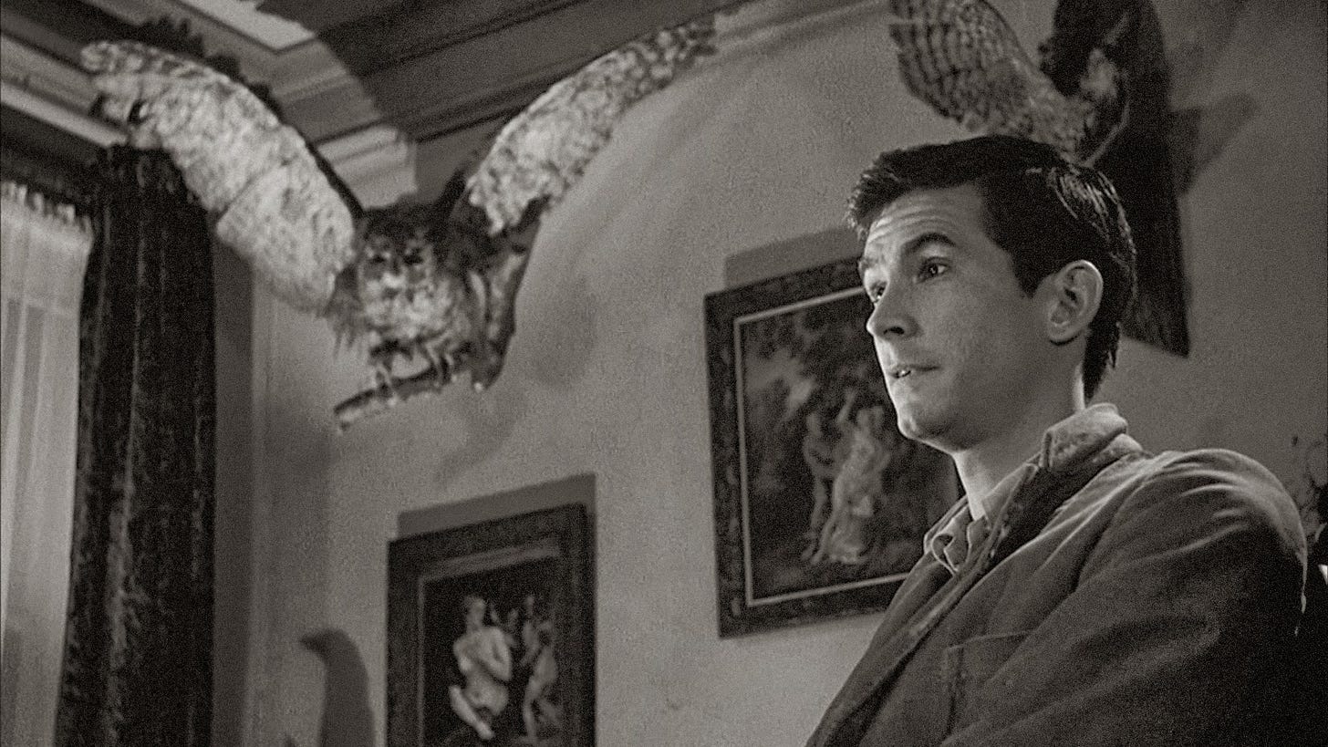 Black-and-white still from Psycho (1960) depicts Norman Bates, played by Anthony Perkins, seated on the right side of the frame with a stuffed owl in the upper left, wings spread as if about to take flight.