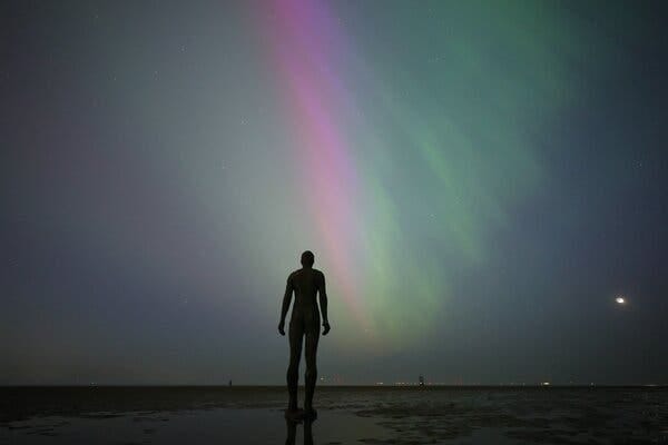A sculpture in human form is visible in front of a night sky filled with purple and green streaks of light.