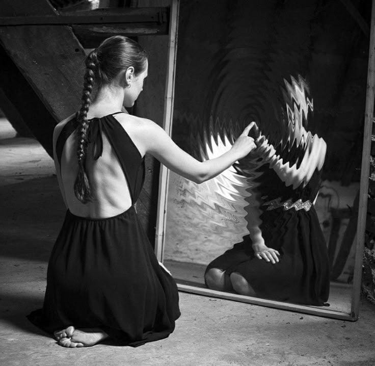 Ta black and white photo of a woman sitting on the ground in front of a mirror with the reflection warping at her touch