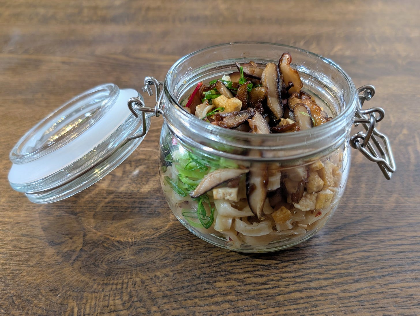 A glass jar with a hinged lid sits open on a wooden surface, filled with neatly layered ingredients: sliced mushrooms on top, a middle layer of chopped green vegetables, and beneath them a mix of noodles and small tofu cubes. The clear jar shows the contrasting textures and colours stacked tightly together.