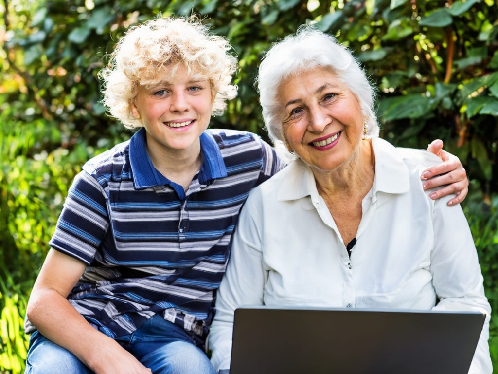 White teenager boy with curly blond hair and blue striped shirt sits with his left arm around his grandmother, who has white hair and white shirt – sitting in a garden setting with laptop in foreground. Both smiling into camera.