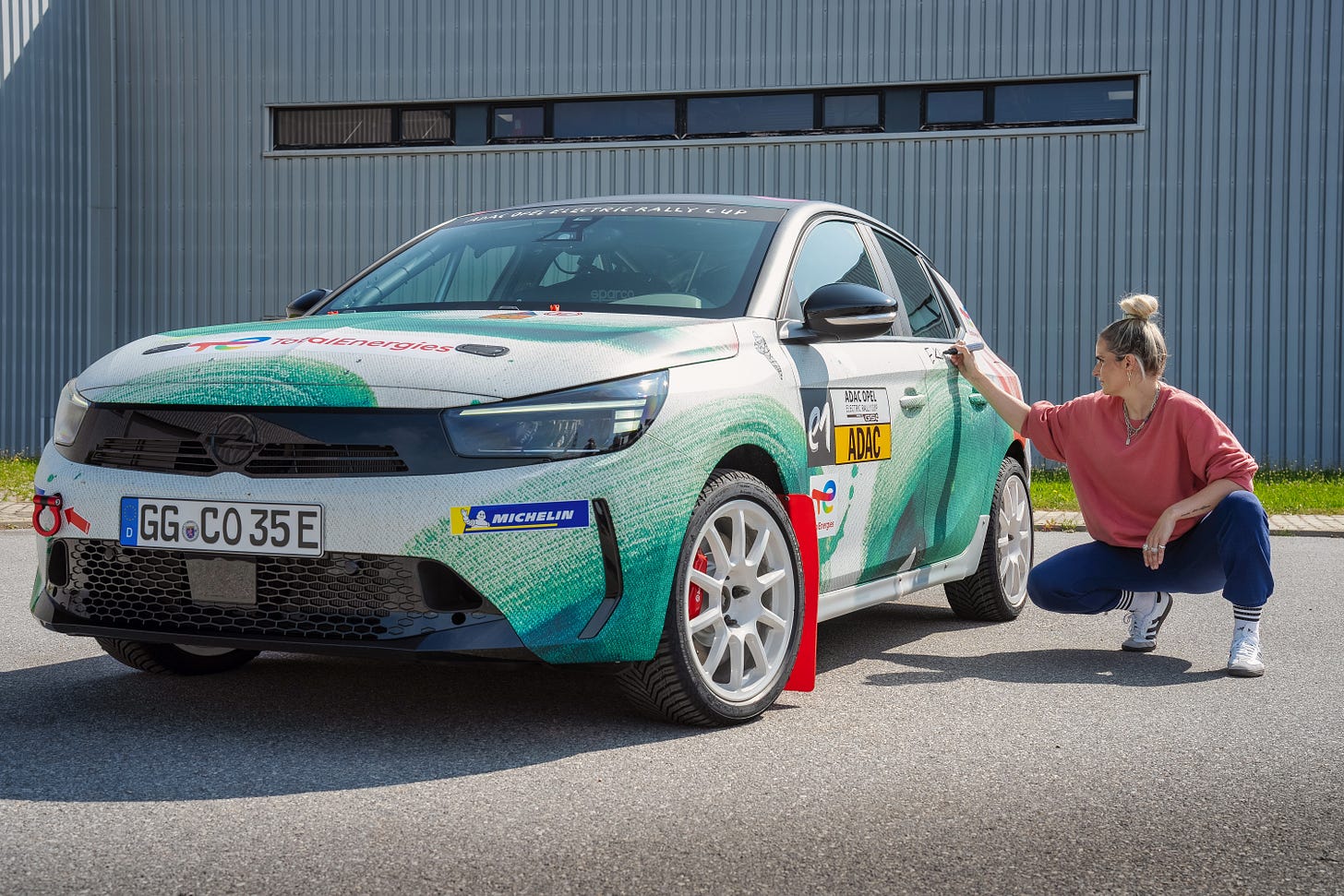 Una foto de un coche con una persona agachada junto a él. El coche es un moderno hatchback con un envoltorio de camuflaje verde y azul. El coche tiene una matrícula que dice “GG-CO35E”. El coche tiene una pegatina roja y blanca en el parachoques delantero que dice “Opel e-Rally Cup”. La persona está agachada junto a la rueda delantera del coche. El fondo es un edificio industrial gris con un cielo azul.