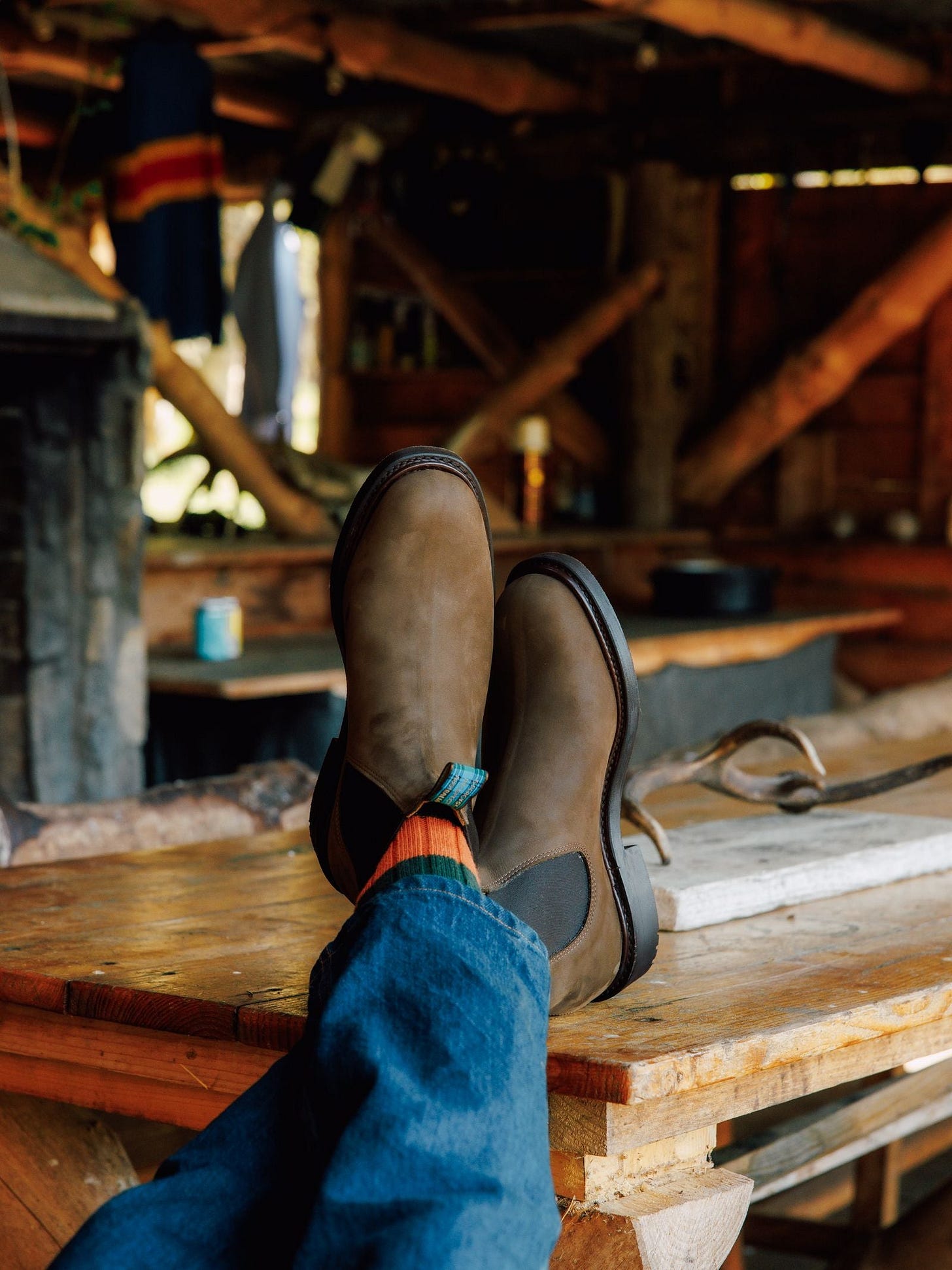 Relaxing in a cozy wooden cabin, a person lounges with feet up on a rustic table, showcasing Men's R.M.Williams Highlander Boots by R.M.Williams x Campbell's of Beauly, paired with orange socks and blue jeans amid wooden beams and hanging clothes. Relaxing in a cozy wooden cabin, a person lounges with feet up on a rustic table, showcasing Men's R.M.Williams Highlander Boots by R.M.Williams x Campbell's of Beauly, paired with orange socks and blue jeans amid wooden beams and hanging clothes.