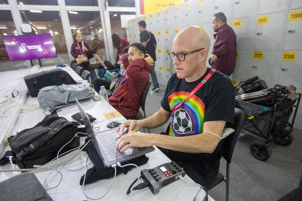 Two men sit at a long table, working on laptops. The man on the right wears a black T-shirt with a soccer ball surrounded by a rainbow. Two men sit at a long table, working on laptops. The man on the right wears a black T-shirt with a soccer ball surrounded by a rainbow.