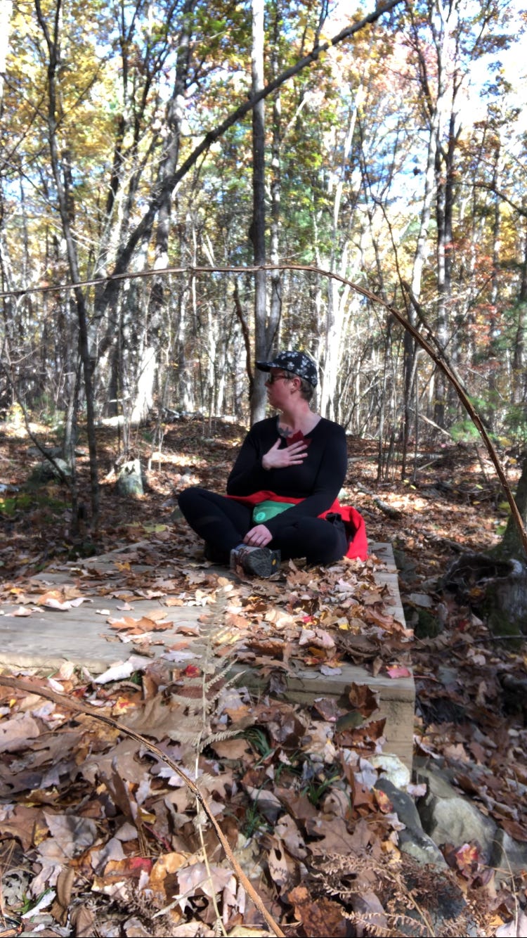 A person dressed mostly in black sits on a small wooden platform in the woods in daylight pressing a leaf to their chest